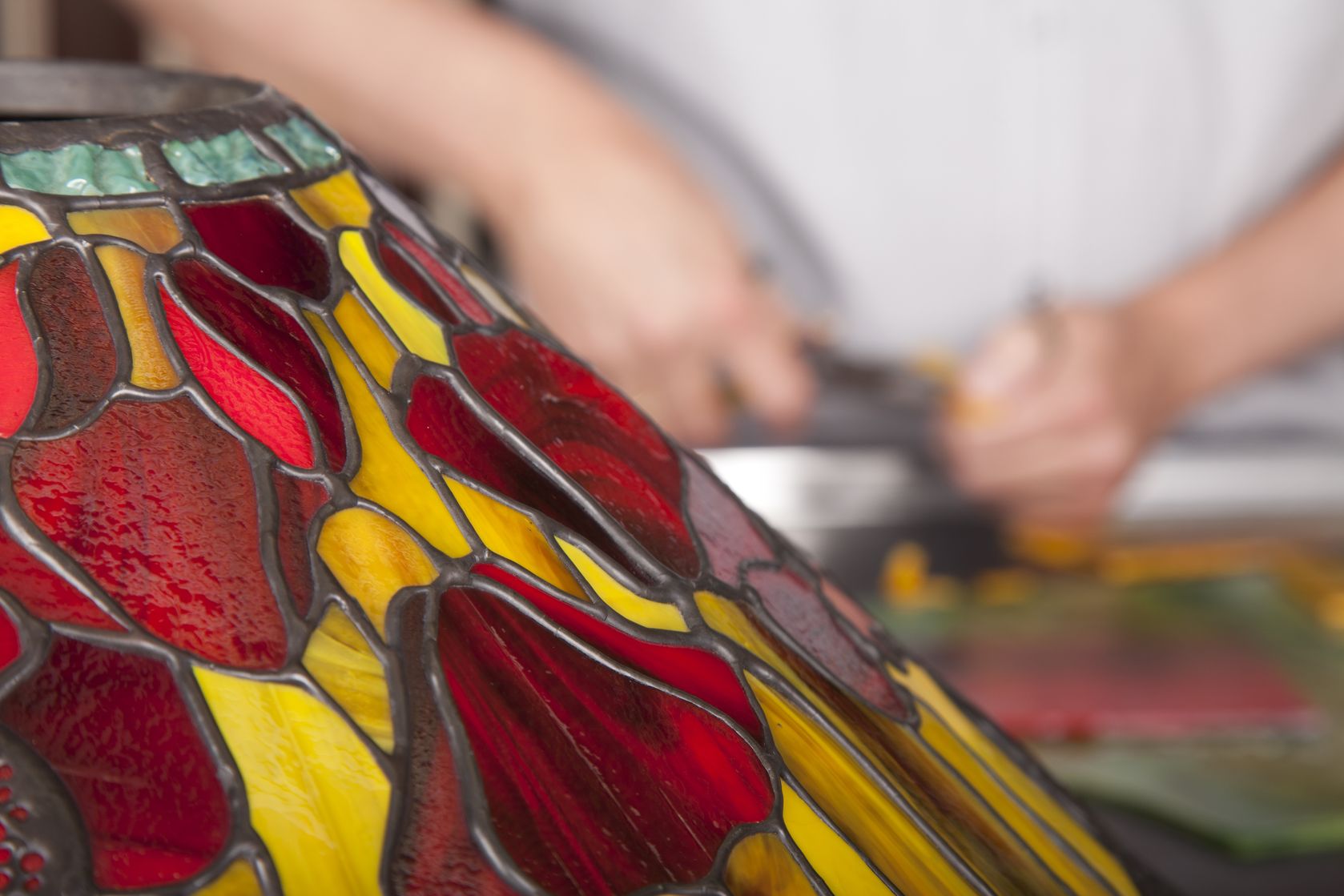 A clean table lamp with stained glass stands in the foreground, while in the background, a person is seen cutting something.