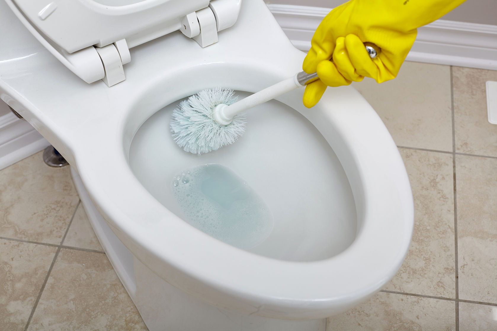 Hand in yellow gloves scrubbing a toilet bowl with a brush to remove hard water stains.