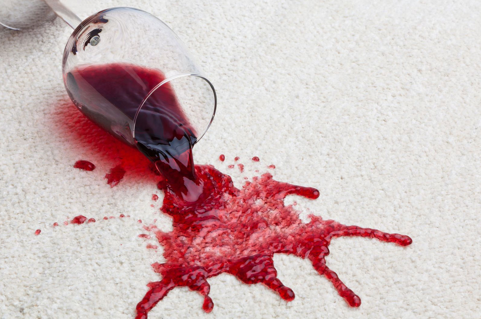 A wine glass tipped over, spilling red wine onto a white carpet in a living room.